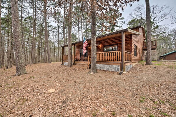 A rustic cabin with a wooden deck, surrounded by tall trees. - Cabin w/ Hot Tub & Grill - 6 Mi to Broken Bow Lake (Broken Bow)
