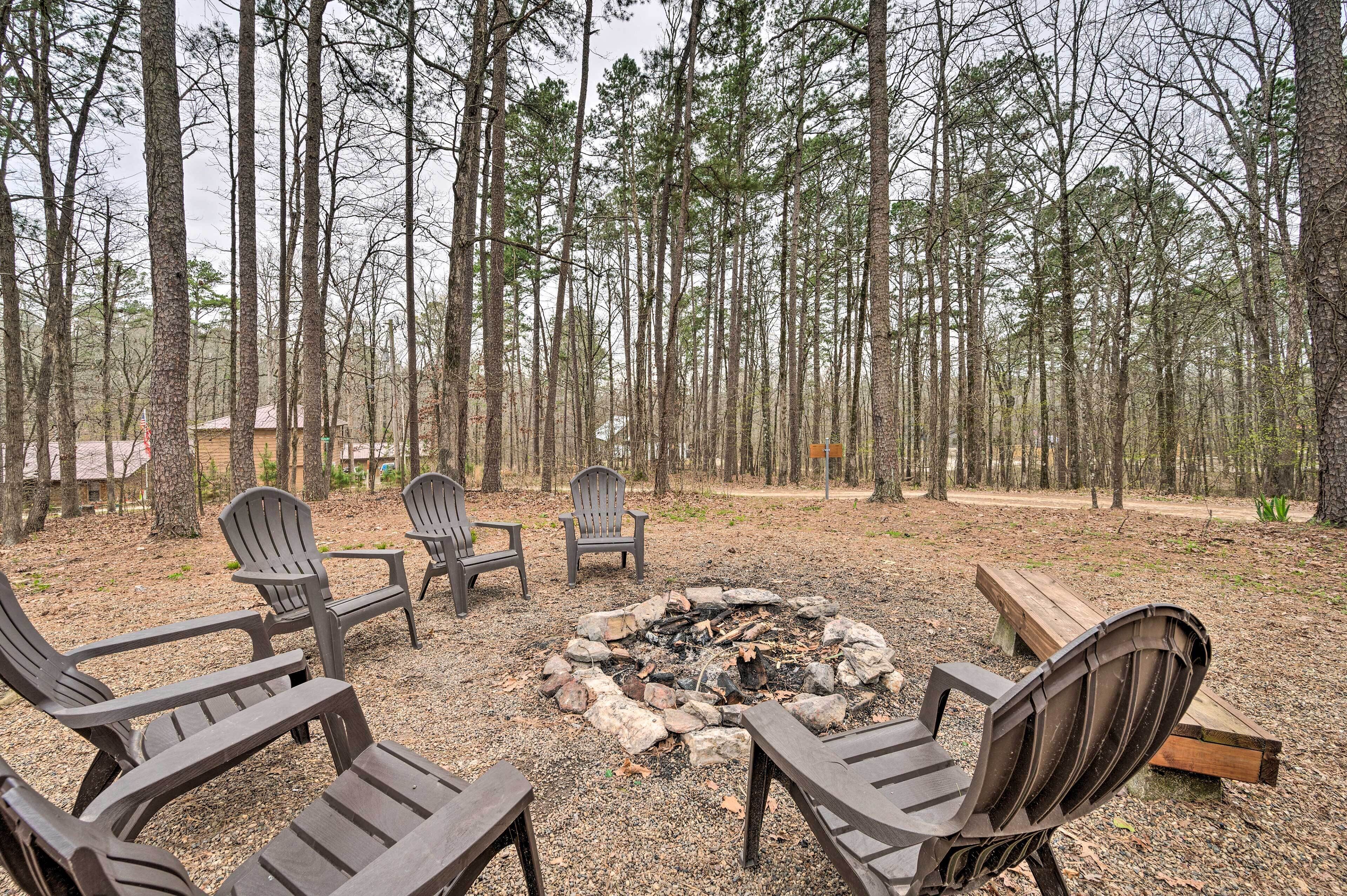 A forested area with a fire pit and Adirondack chairs.