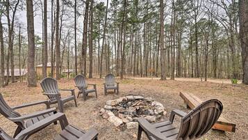 A forested area with a fire pit and Adirondack chairs.