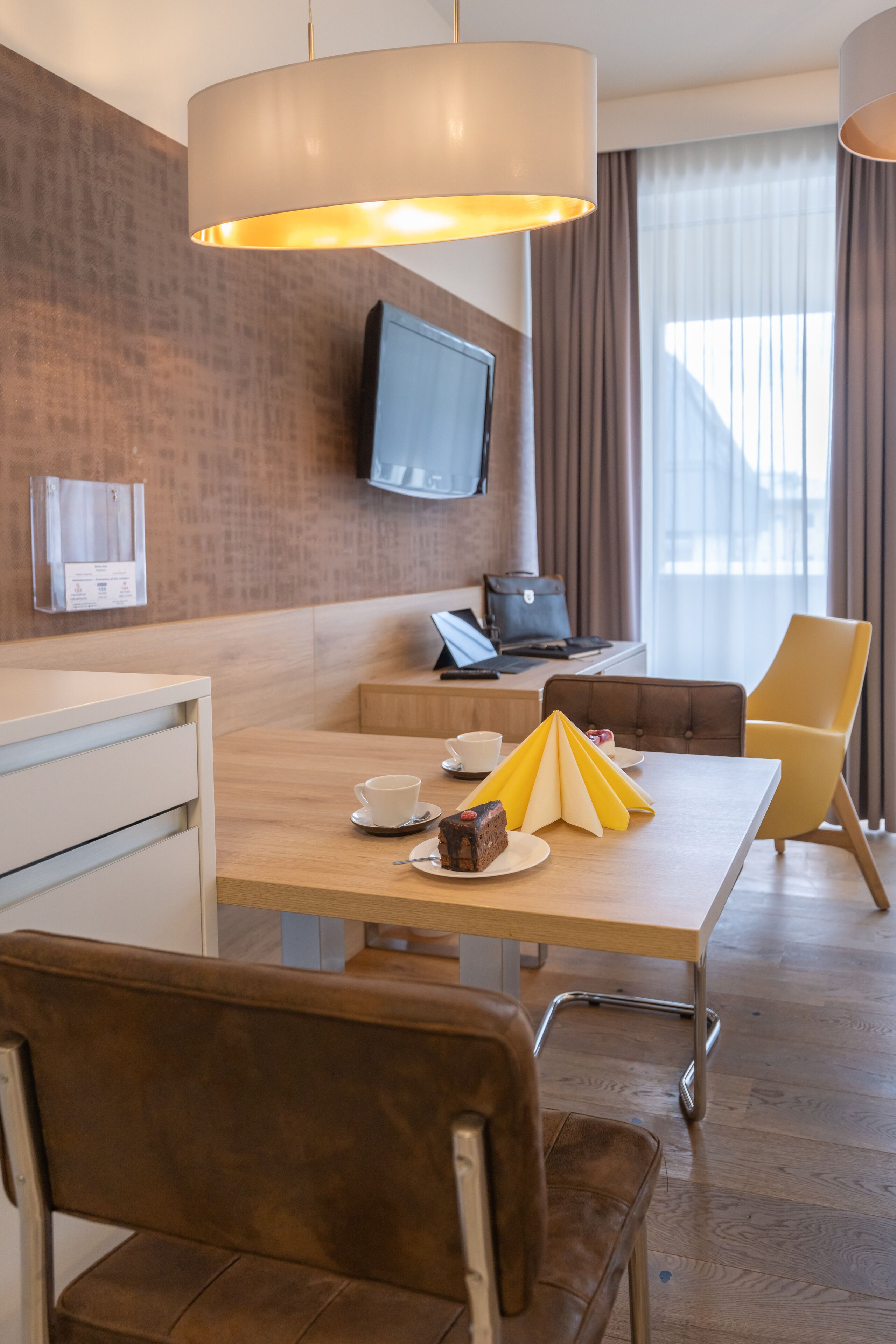 A hotel room with a wooden desk, a television mounted on the wall, a laptop, a yellow chair, and a brown leather chair.