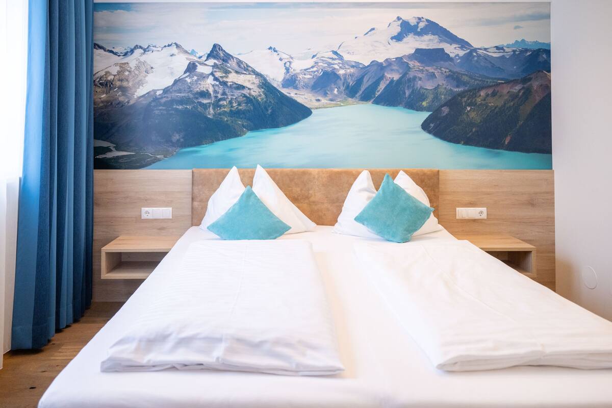 Habitación de hotel con una cama, dos almohadas, cabecera de madera, mesita de noche y un ventanal con cortinas.