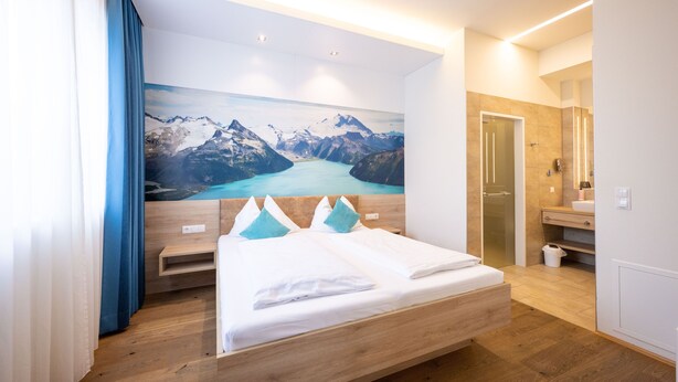 A hotel room with a wooden bed, a large mountain landscape painting, a wooden nightstand, and a bathroom with a mirror and sink. - PM PremiumAPART Strassgang (Graz)
