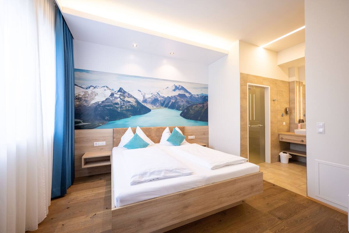 Habitación de hotel con cama de madera, un cuadro de un amplio paisaje montañoso, mesita de noche de madera y baño con espejo y lavamanos.