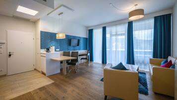 Habitación de hotel moderna con suelo de madera, zona de comedor con mesa y sillas, sofá, televisor montado en la pared y ventanas amplias con cortinas azules.