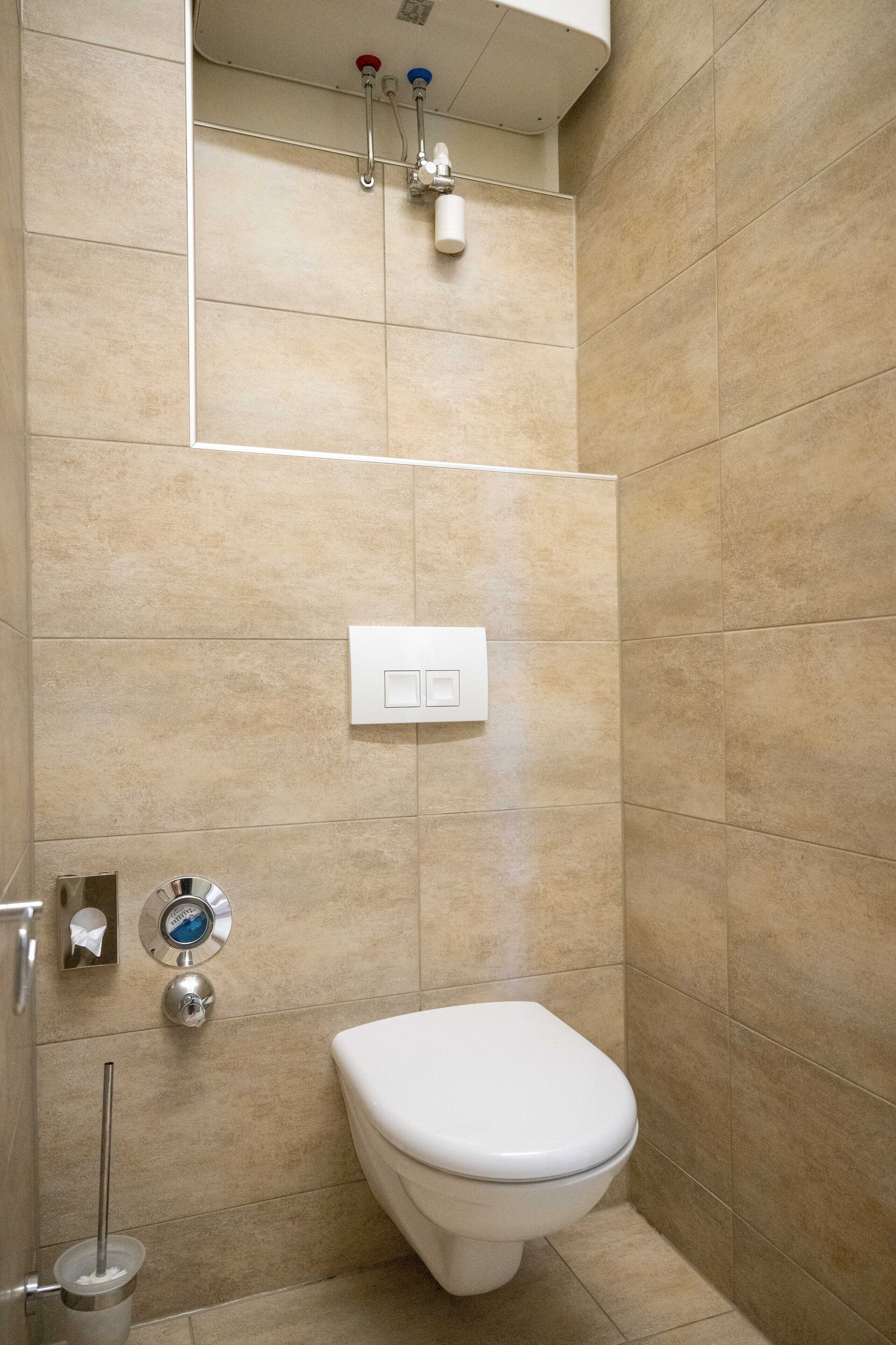 Un baño con azulejos color beige, un inodoro blanco, un secador de manos y un grifo empotrado en la pared.