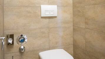 Un baño con azulejos color beige, un inodoro blanco, un secador de manos y un grifo empotrado en la pared.
