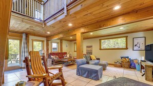 A spacious living room with a high ceiling, wooden flooring, and large windows. There is a wooden rocking chair, a blue sofa, and a wooden coffee table. - Saranac Lake Cabin w/ Deck: Pets Welcome! (Saranac Lake)