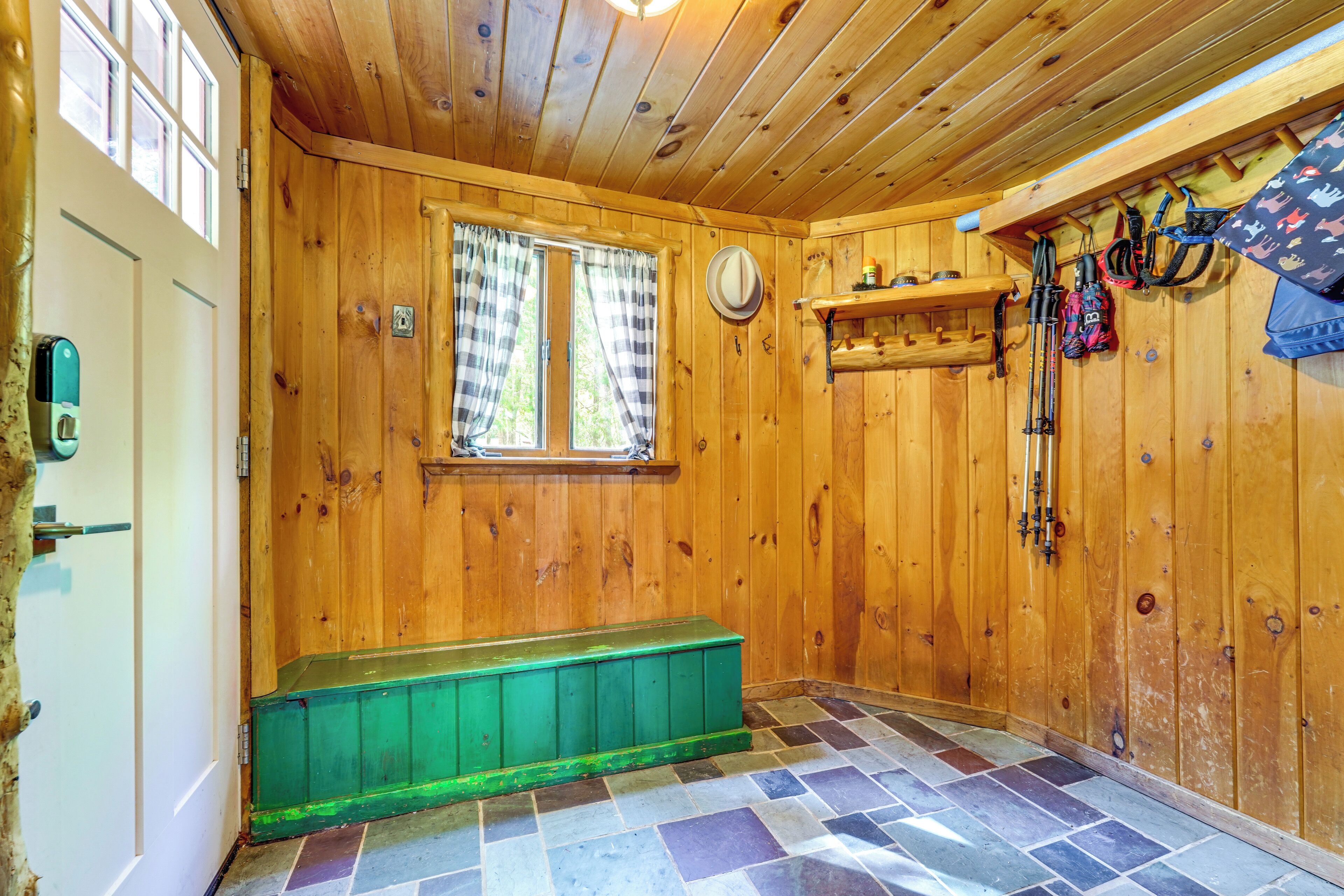 Interior de una cabaña de madera con un banco, una ventana con cortinas y equipo de esquí en la pared.