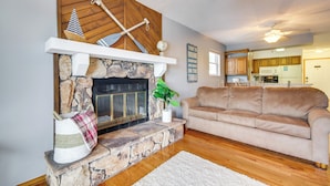 A living room with a stone fireplace, a beige sofa, and a kitchen in the background. - Cozy Lake Ozark Condo Rental w/ Grill, Pool Access (Lake Ozark)