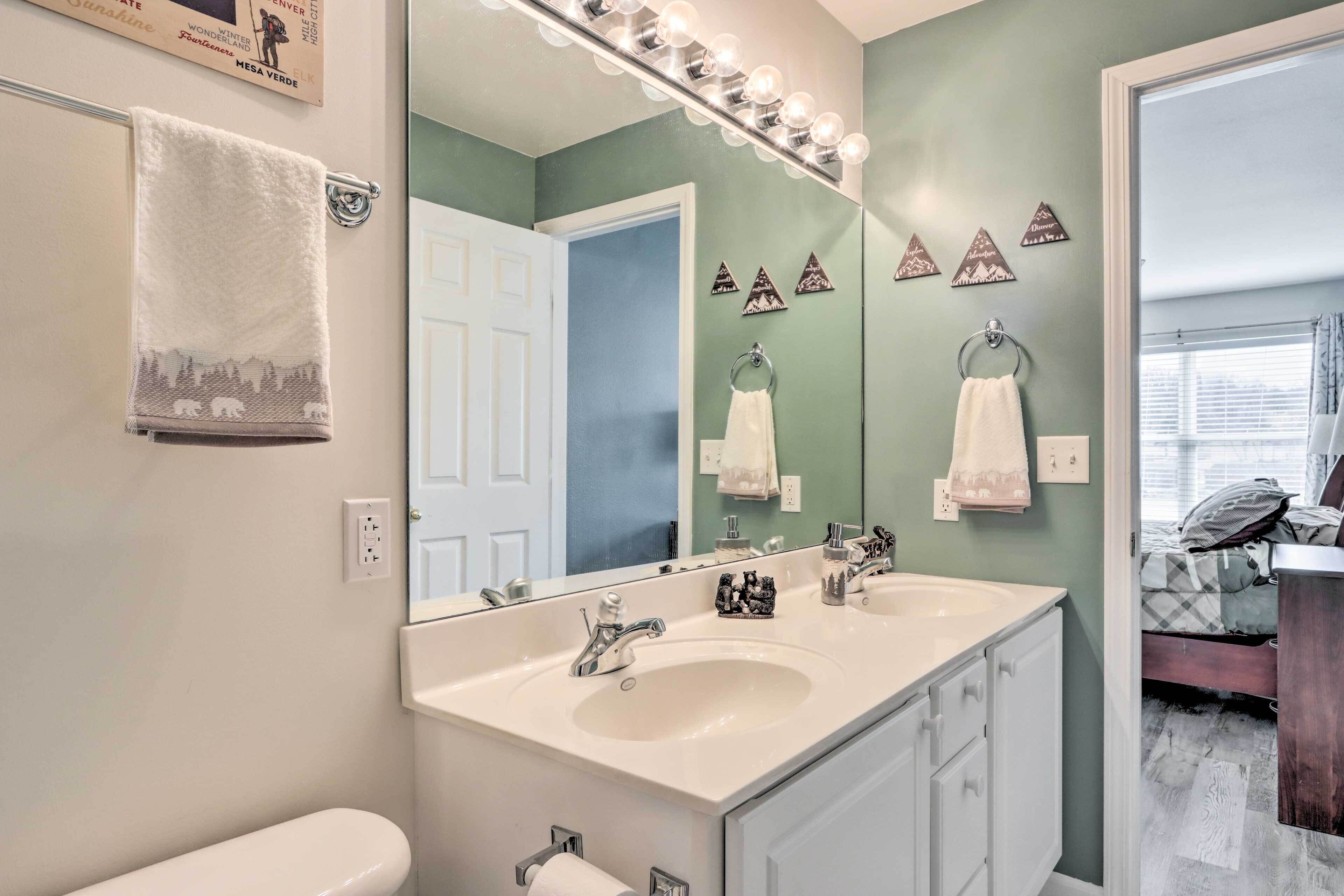 A bathroom with a large mirror, two sinks, and a toilet. There are decorative wall hangings and a towel rack.