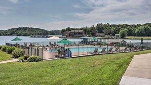 Zona de piscina con sillones de descanso y sombrillas, rodeada por una valla, con un lago y edificios al fondo.