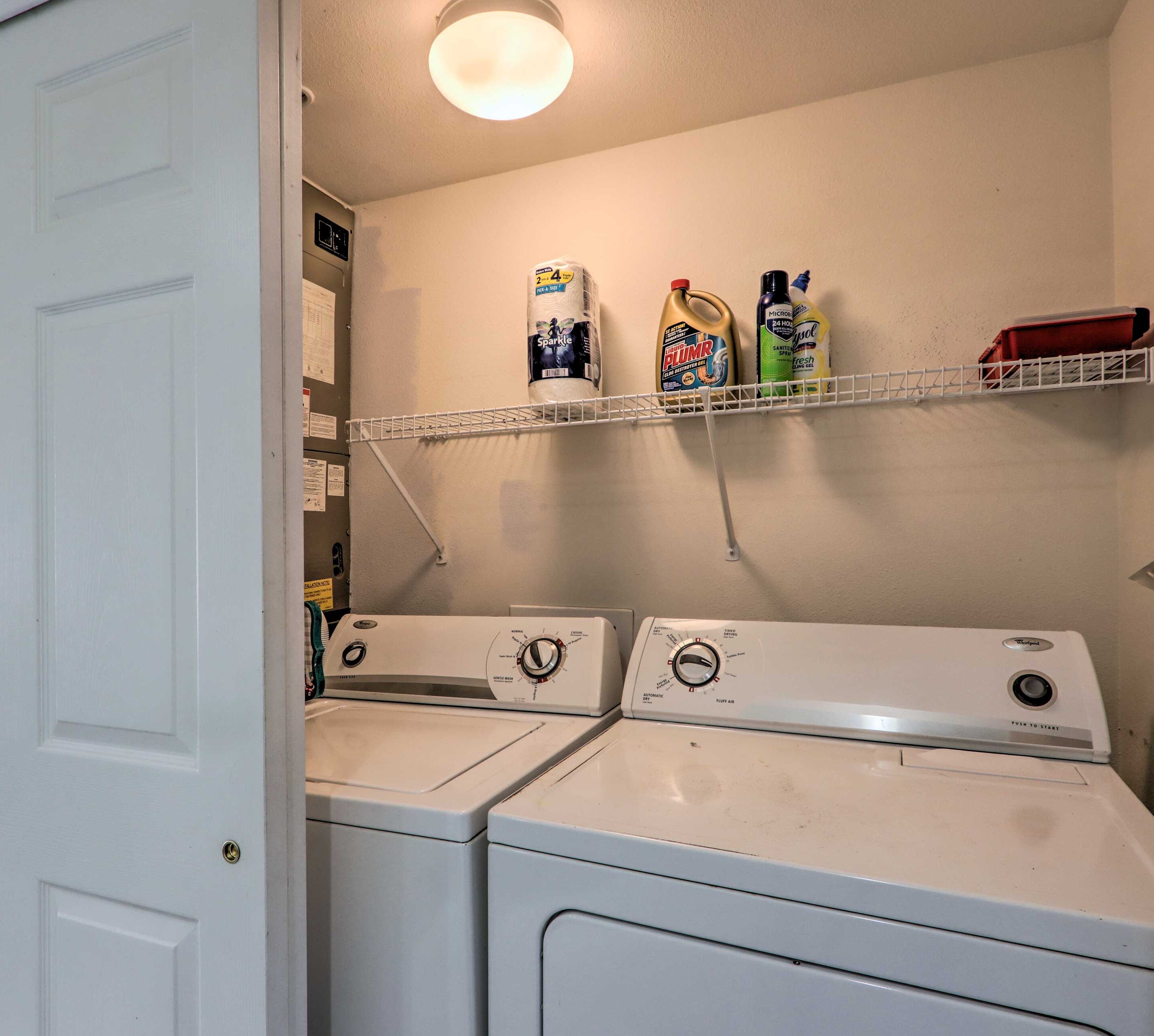 A laundry room with two front-loading washers and dryers, a shelf with cleaning supplies, and a door leading to another room.