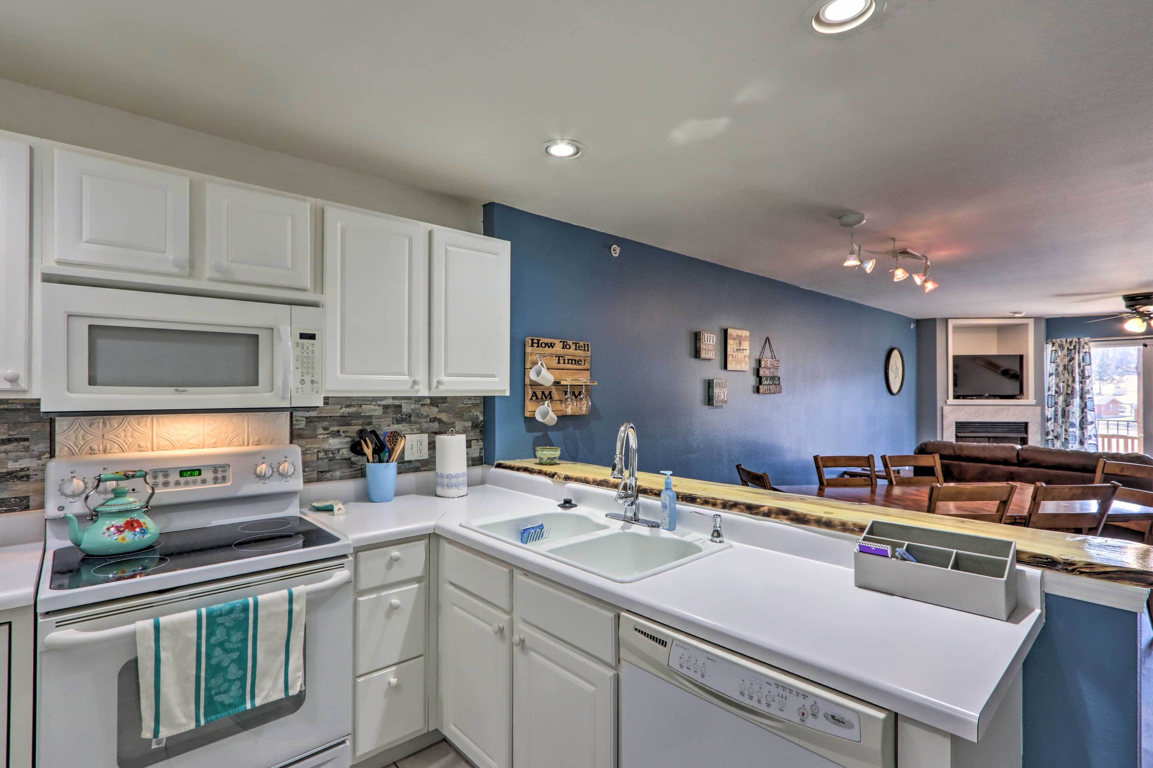 A modern kitchen with white cabinetry, a microwave, an oven, and a dishwasher. There is a blue wall with decorative items and a dining area with a brown leather couch.