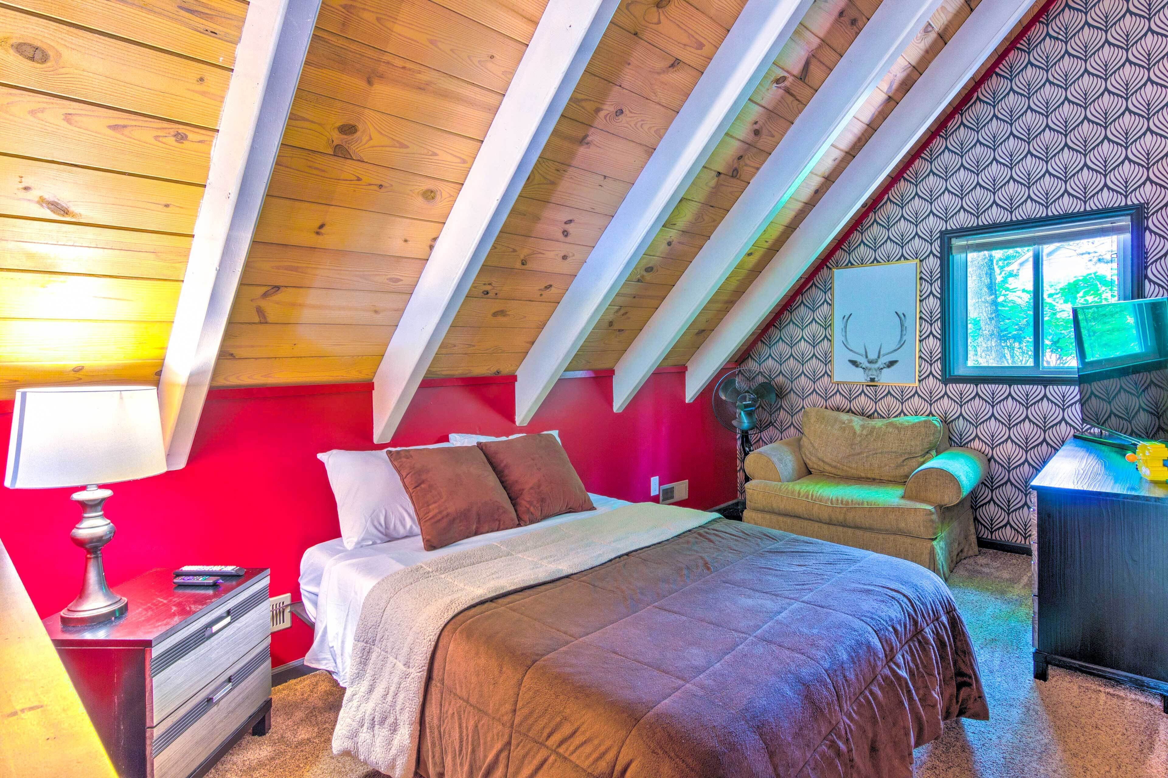 A bedroom with a sloped ceiling, a bed with a brown blanket, a chair, a desk, and a window with a view of trees.