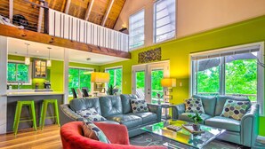 A spacious living area with a high ceiling, a red armchair, two grey sofas with patterned cushions, a glass coffee table, and a kitchen area with a bar counter and stools. - Mtn-view Gem in Lake Lure w/ Game Room & Fire Pit (Lake Lure)