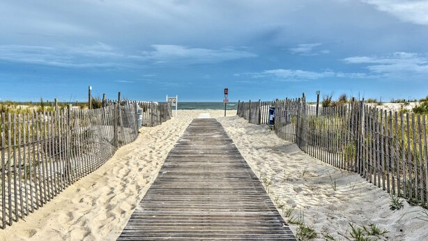 Ein hölzerner Steg führt zu einem Strand, beidseits begrenzt von einer Leitplanke.