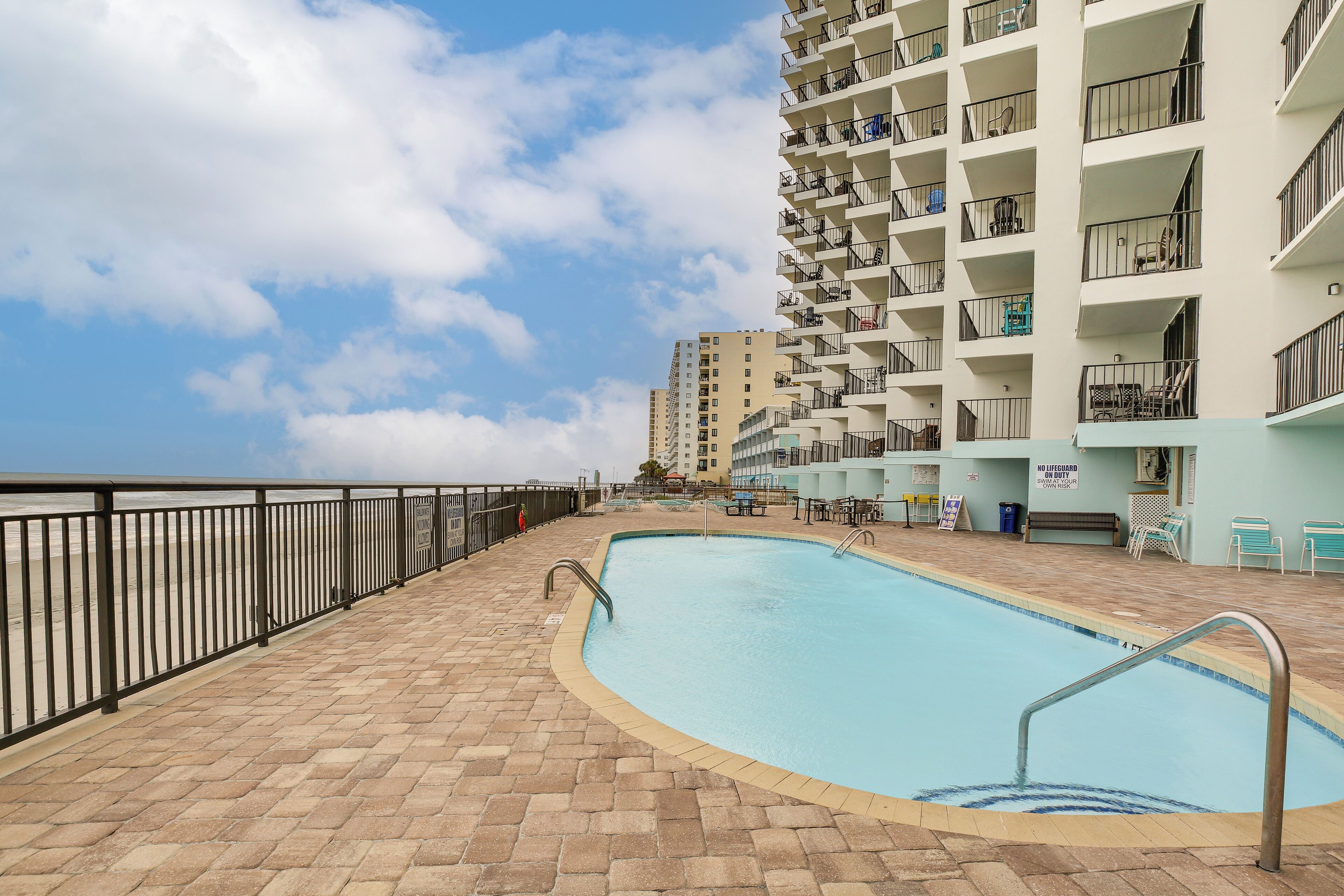 A swimming pool with a tiled deck, a metal railing, and a view of a high-rise building with balconies.