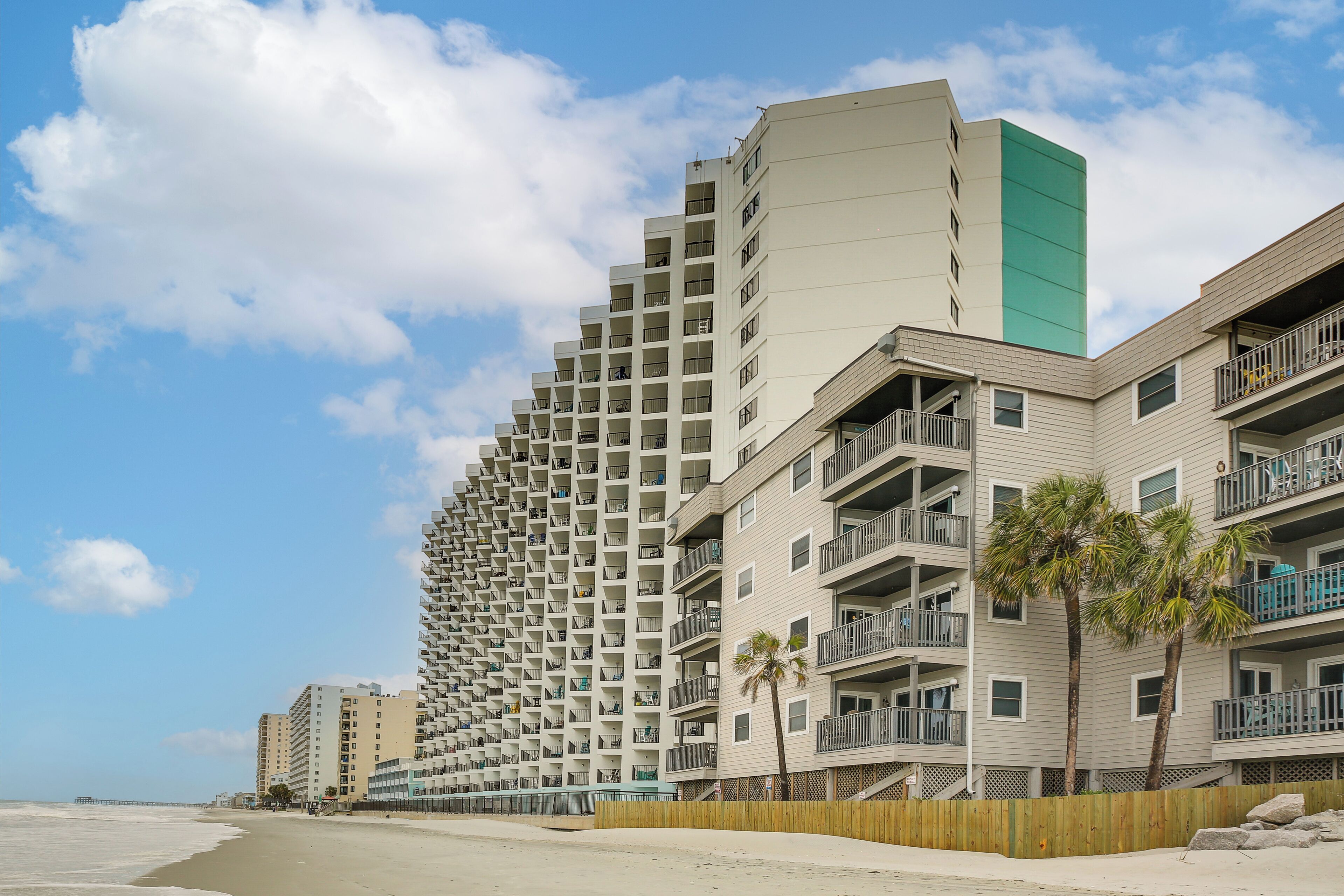 A beachfront hotel with multiple balconies, a wooden fence, and palm trees.