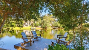 A lakeside deck with Adirondack chairs and a dock extending into a calm lake, surrounded by lush greenery and a distant building. - Charming Lakefront Retreat in Palmetto Dunes! (Hilton Head Island)