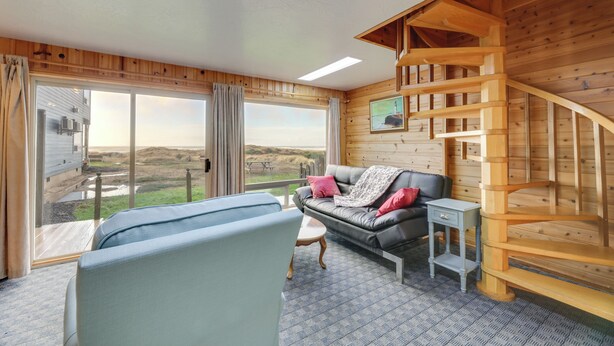 A room with a large window, a sofa, a small table, and a wooden staircase. - Sanderling Sea Cottages, Unit 3 w/ Private Balcony (Waldport)