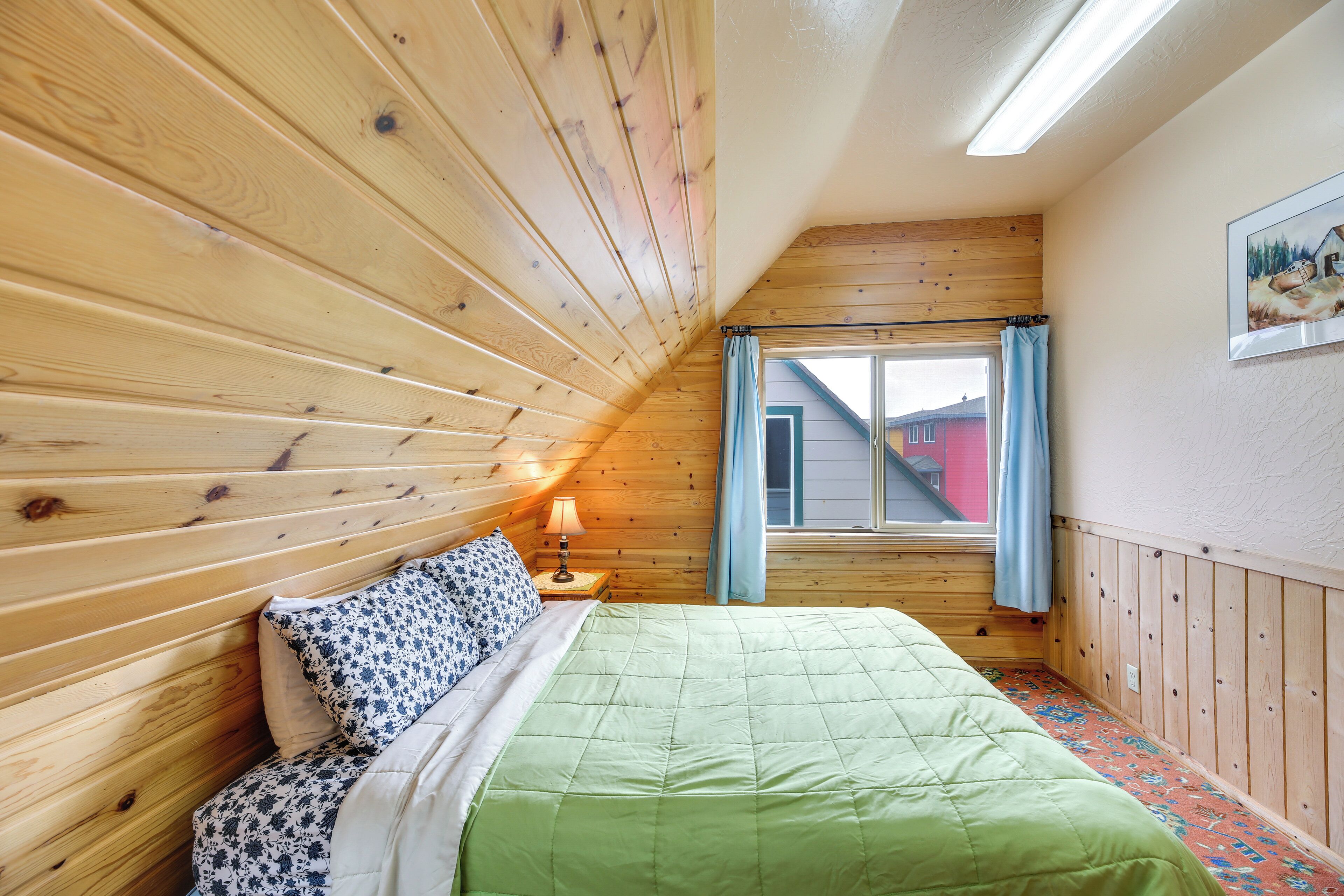 A room with wooden paneling, a bed with green bedding, a nightstand with a lamp, and a window with curtains.