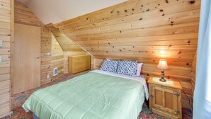 A cozy bedroom with a wooden sloped ceiling, a bed with a green quilt, a nightstand with a lamp, and a wooden wardrobe. - Sanderling Sea Cottages, Unit 3 w/ Private Balcony (Waldport)