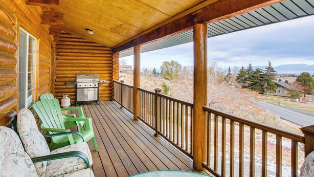 A wooden cabin with a porch, green Adirondack chairs, a grill, and a view of a road and trees.