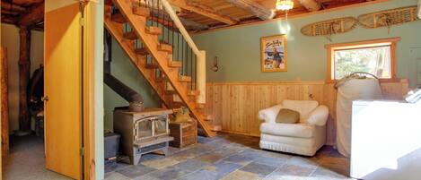 A rustic interior with a stone-tiled floor, a wooden staircase, a white armchair, and a vintage stove.