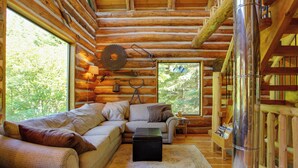 A cozy log cabin interior with a sofa, ottoman, and a large window offering a view of the forest. - Cozy Easton Cabin w/ Wenatchee Nat'l Forest Views! (Easton)