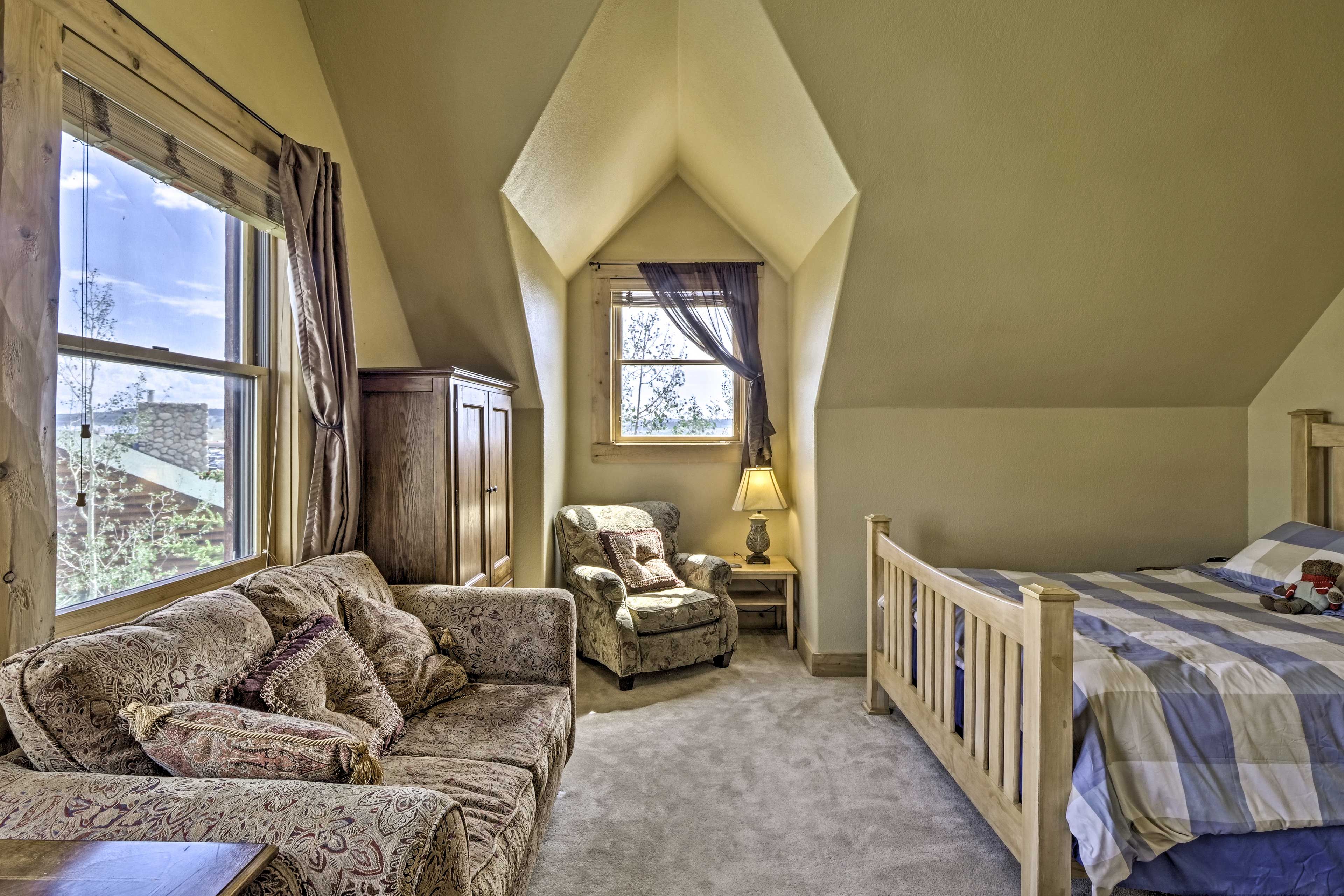 A cozy bedroom with a sloped ceiling, a large window with curtains, a patterned sofa, a chair, a bedside table with a lamp, and a bed with a checkered bedspread.
