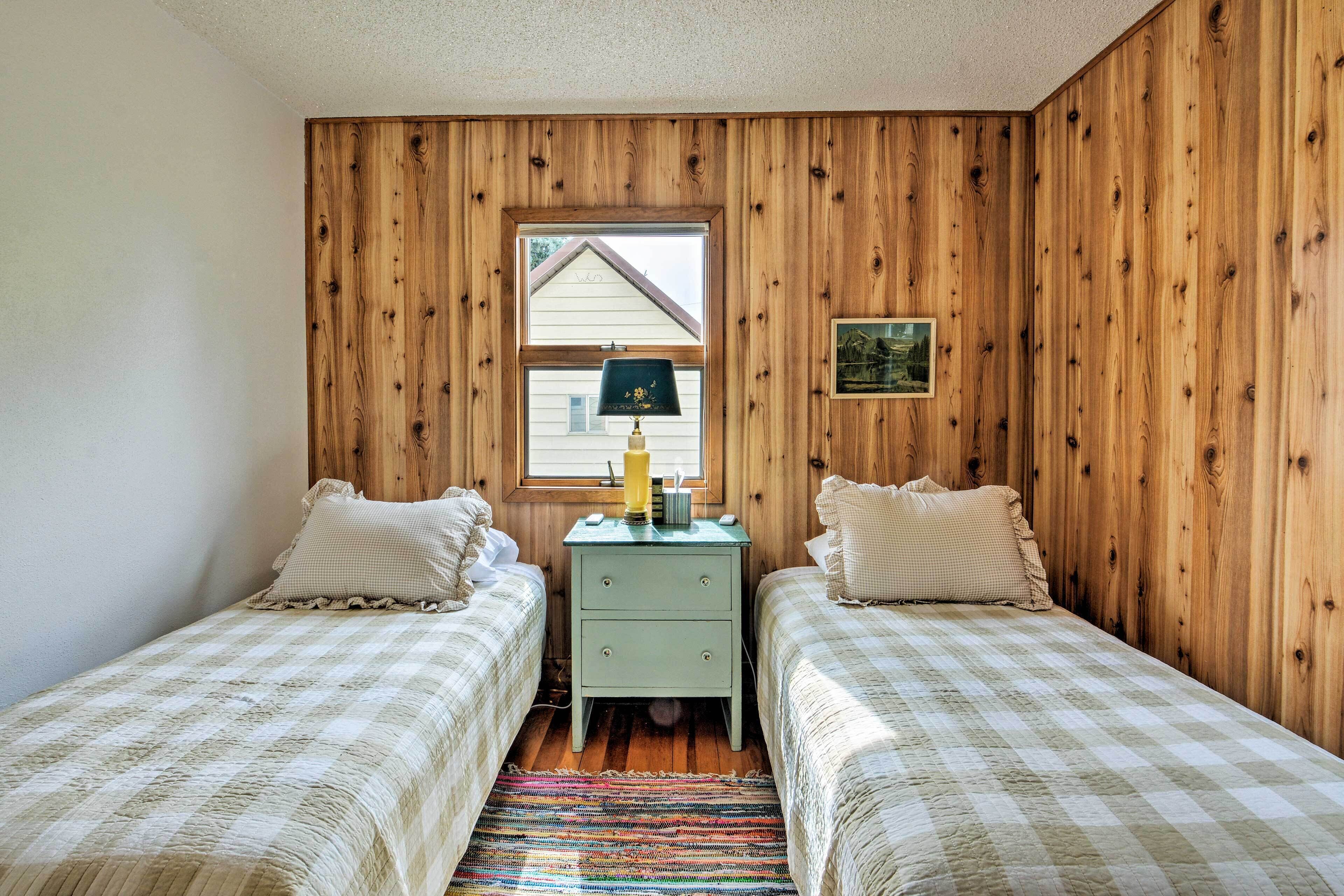 Een kamer met twee eenpersoonsbedden, een klein tafeltje met een lamp, een raam met uitzicht op een huis en houten paneelwanden.