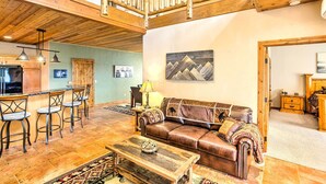 Un amplio salón con techo alto, una escalera de madera, un sofá de cuero marrón, una mesa de centro y una zona de bar con taburetes.