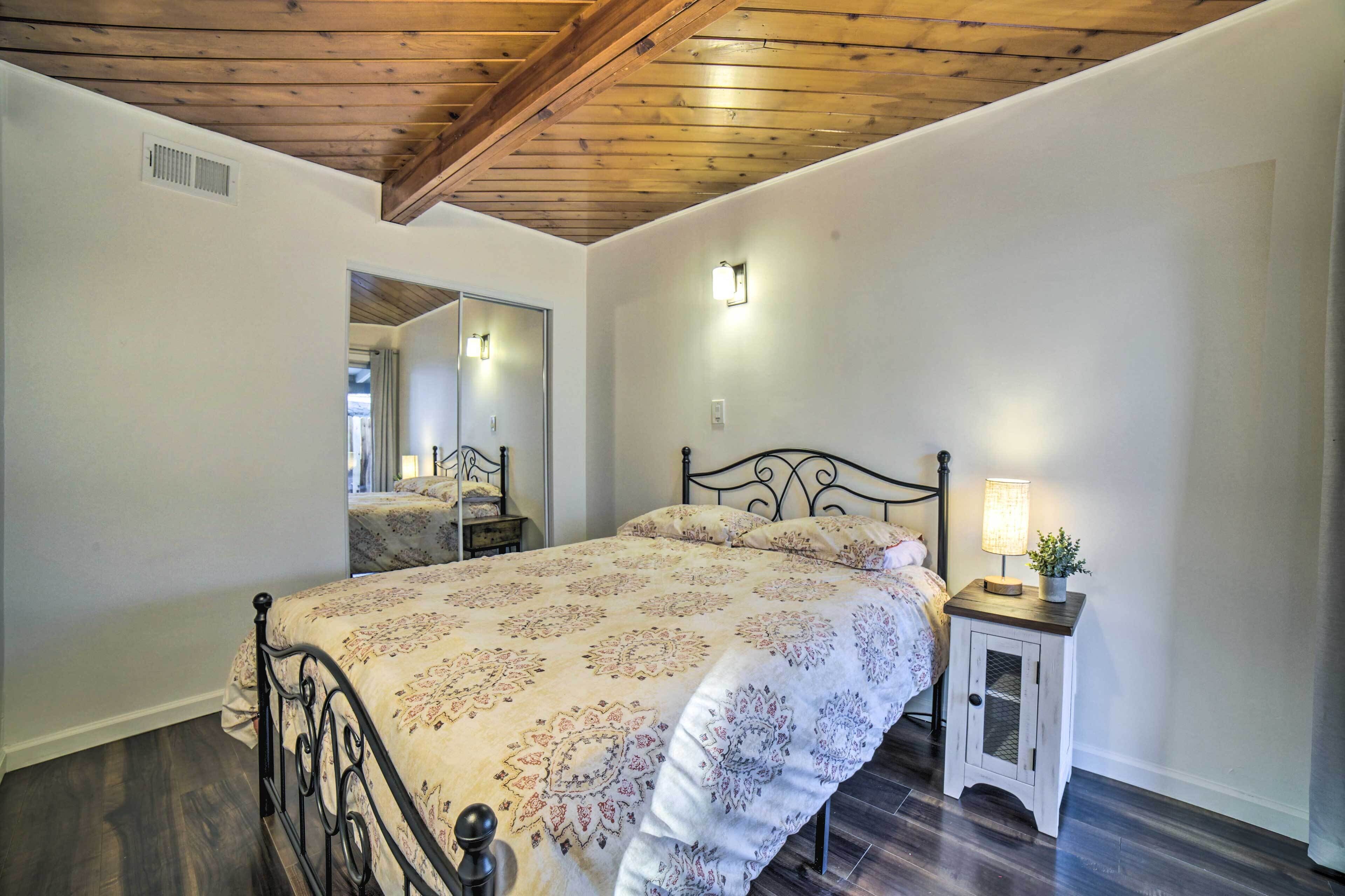 A bedroom with a bed, a nightstand with a lamp, a mirror, and wooden ceiling beams.
