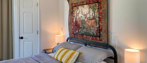 A neatly made bed with a striped pillow, a tapestry with deer on the wall, and a bedside lamp on a wooden nightstand.