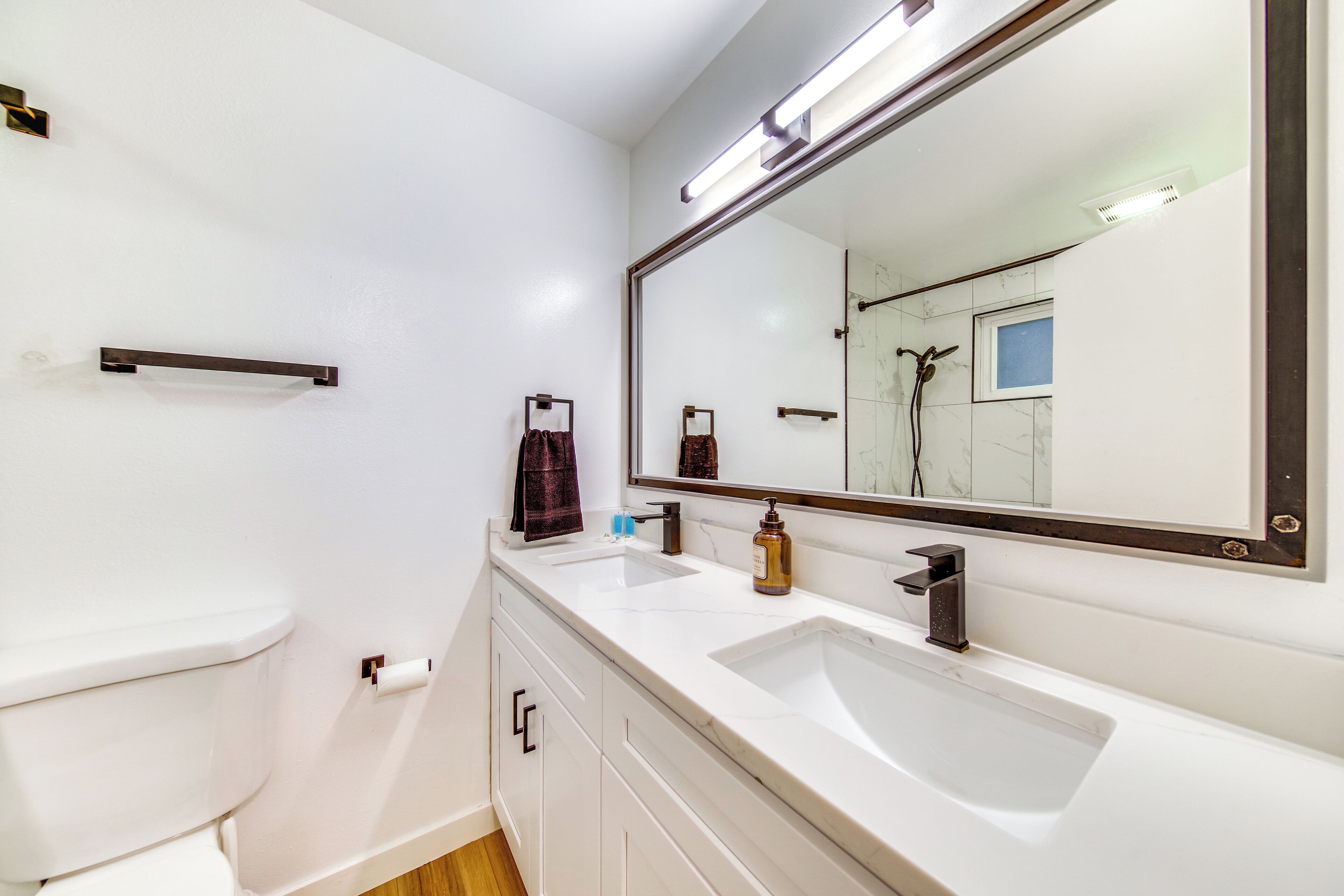 Un baño moderno con inodoro blanco, dos lavamanos, un espejo grande y un perchero para toallas.