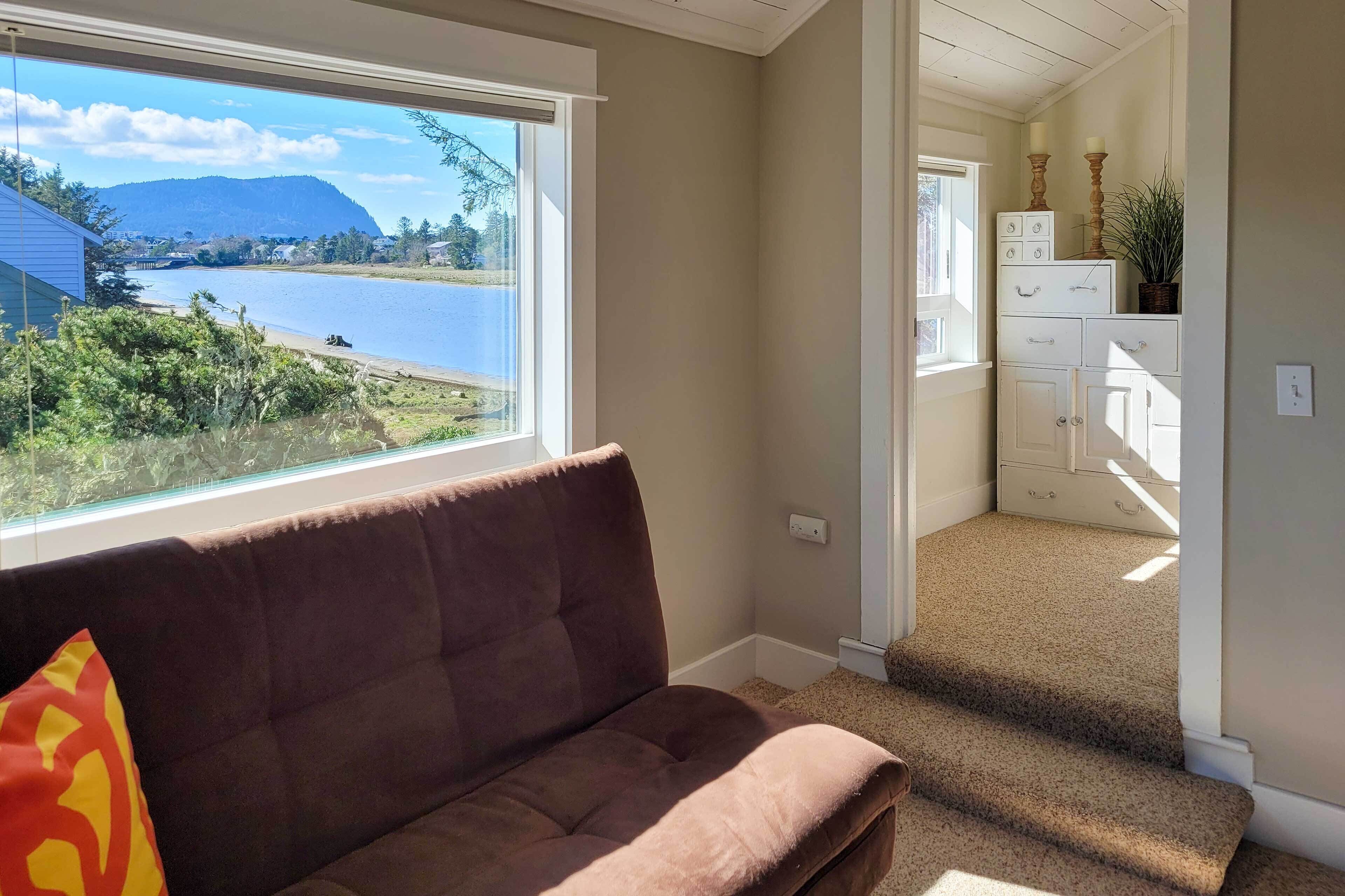 A living room with a brown couch, a window with a view of a lake and mountains, and a staircase leading to a bedroom.