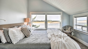 Un dormitorio con una cama grande, una silla y vistas a un cuerpo de agua a través de amplias ventanas.