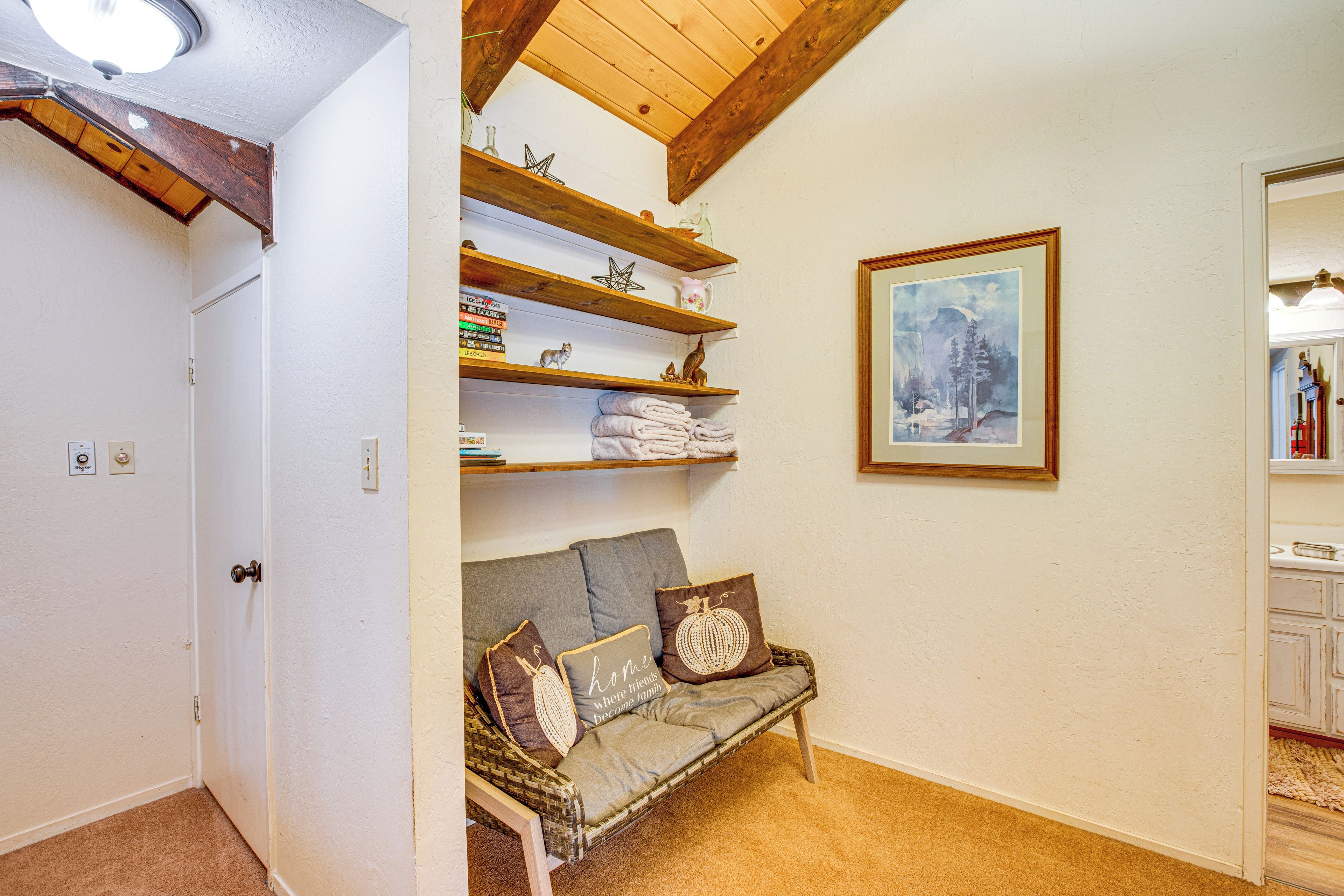Ein gemütlicher Raum mit Holzbalkendecke, einer gepolsterten Bank mit dekorativen Kissen, einem an der Wand befestigten Regal mit Büchern und Dekoration sowie einem gerahmten Gemälde.