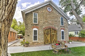 A stone house with a wooden garage door, a small porch with chairs, and a cart with flowers.