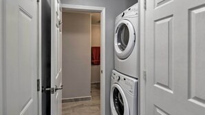 A laundry room with two stacked front-loading washing machines, a door leading to a hallway, and a red towel hanging on a rack. - Charming Historic Condo - Walk to Downtown & UW (Laramie)