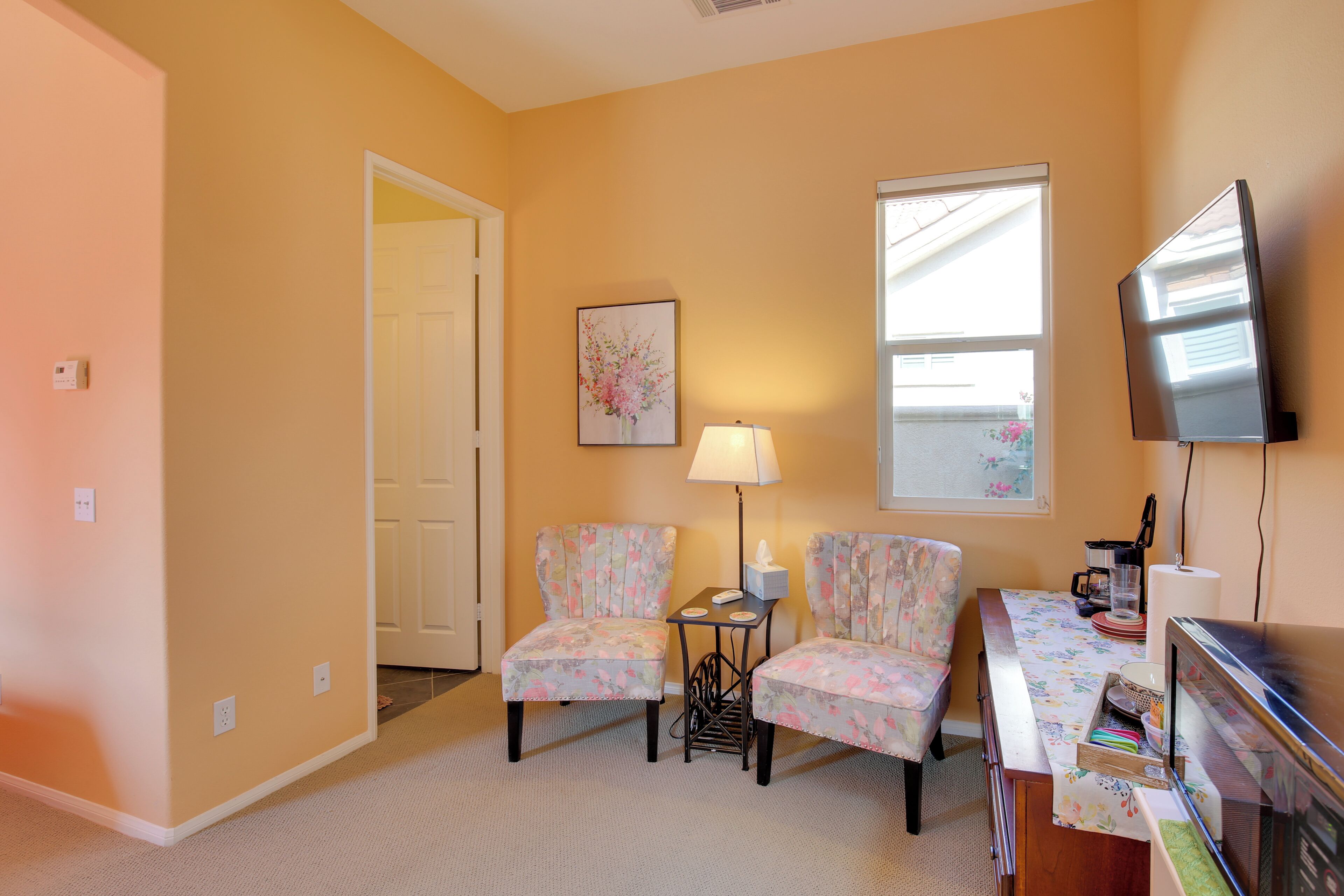 A room with two floral chairs, a small table with a lamp, a television, and a window with blinds.