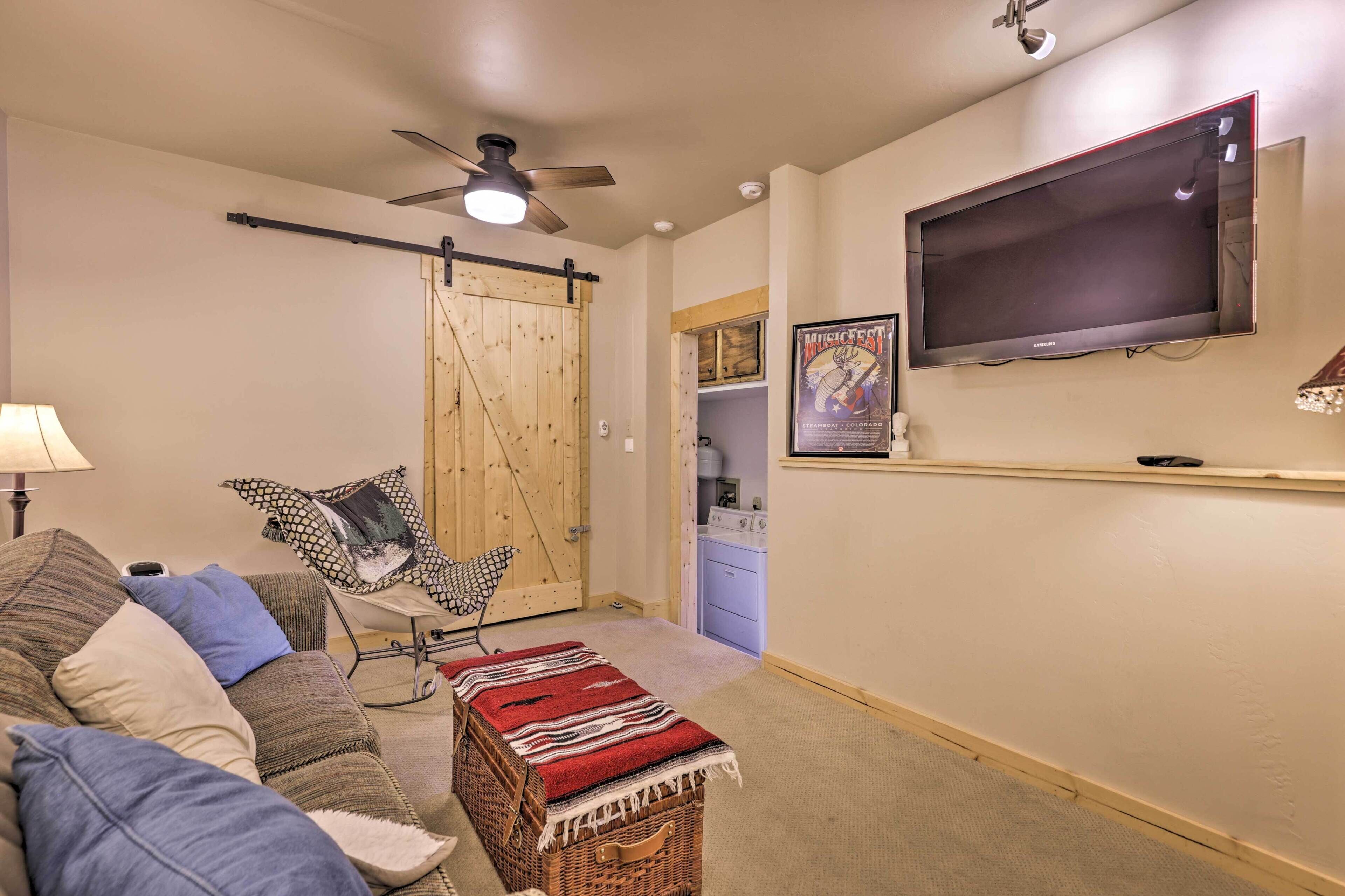 A living room with a sofa, a coffee table, a ceiling fan, a TV mounted on the wall, and a door leading to another room.