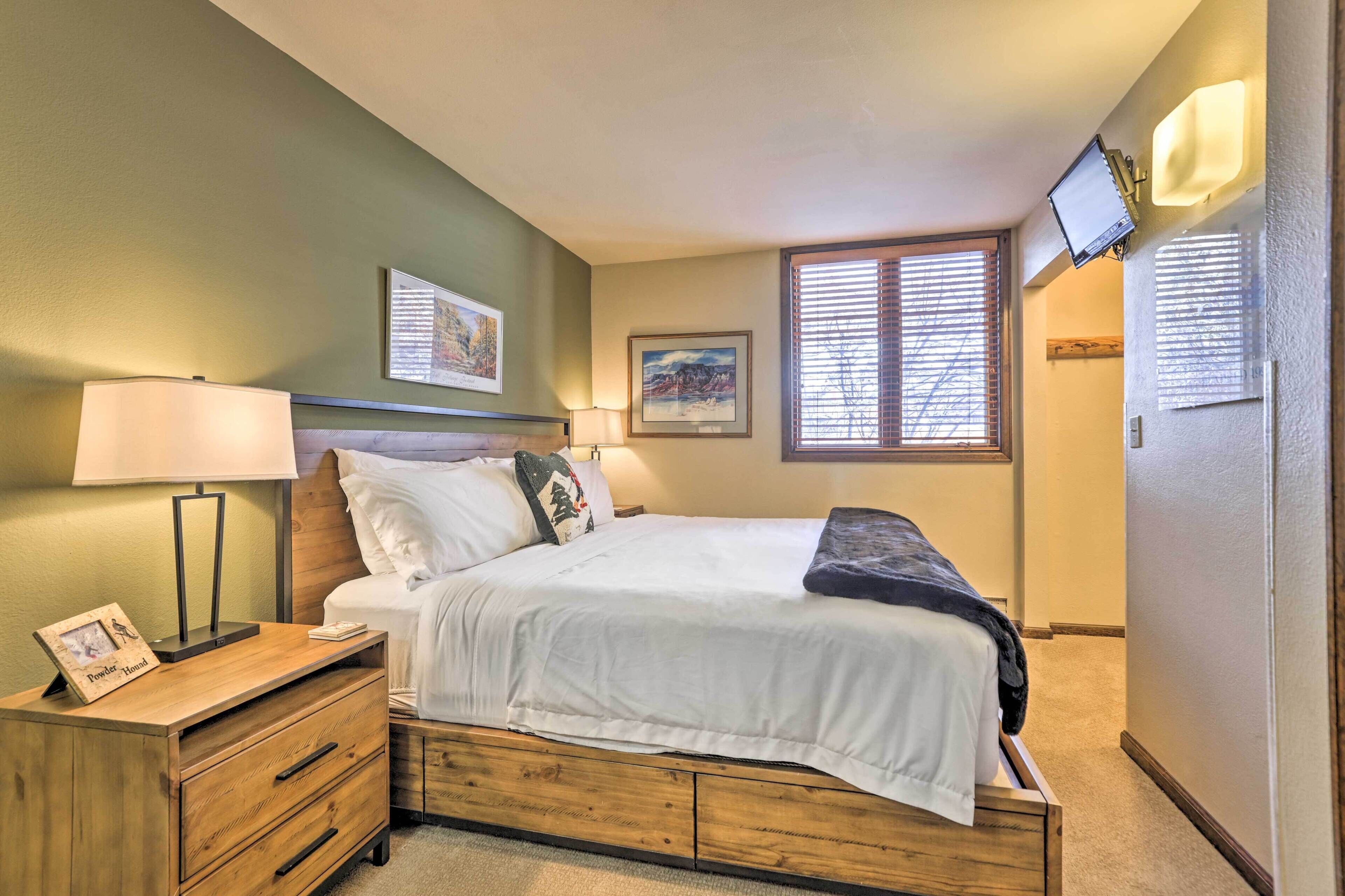 A bedroom with a bed, bedside tables, a TV mounted on the wall, and a window with blinds.