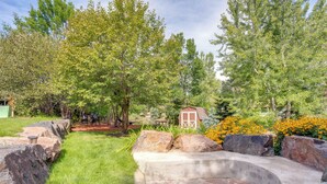 A stone fire pit surrounded by rocks and grass, with trees and a small shed in the background.