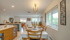 A dining area with a large table, chairs, and a chandelier. There is a kitchen area with wooden cabinets and a sink.