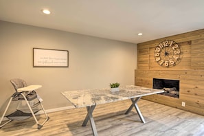 A room with a wooden wall, a large clock, a framed quote, a high chair, and a table with a plant.