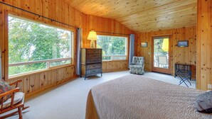 Ein Zimmer mit Holzwänden, einem Bett, einem Schminktisch, einem Sessel und großen Fenstern mit Blick auf Bäume.