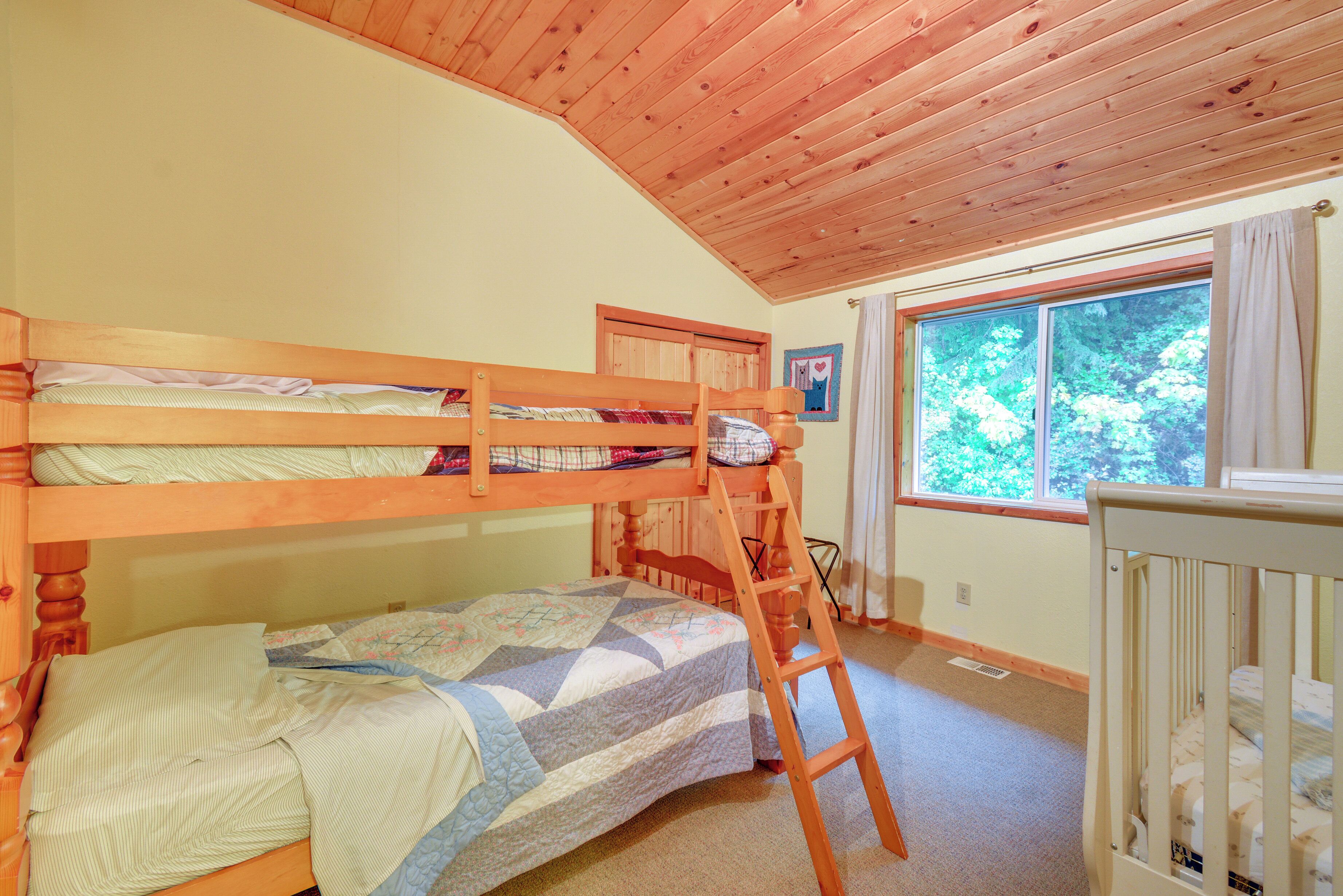 A bunk bed room with a wooden ladder, a crib, and a window with a view of trees.
