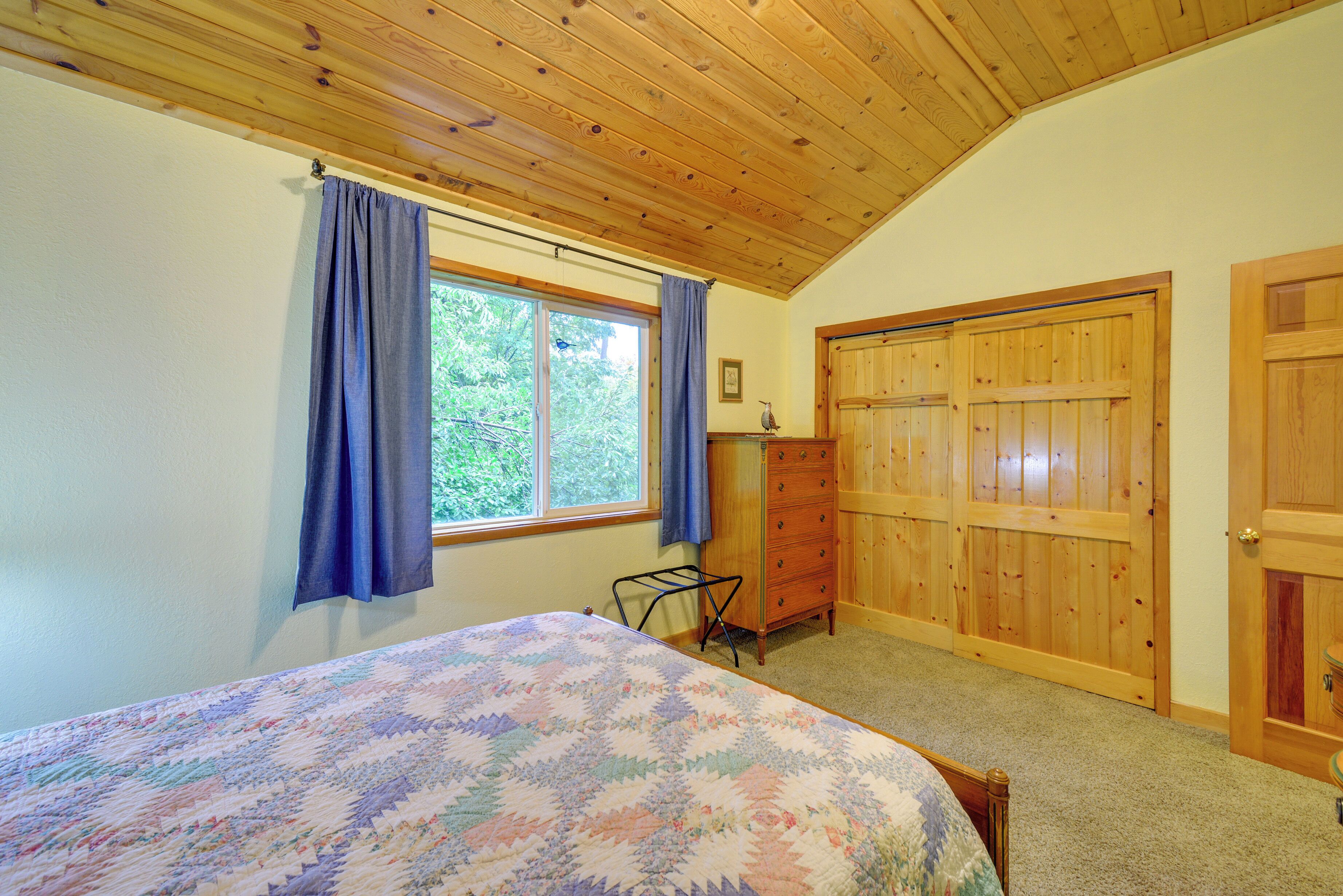 A bedroom with a wooden ceiling, a bed with a floral quilt, a dresser, and a window with curtains.