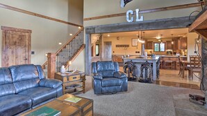 A spacious living room with a leather sofa, a leather armchair, and a coffee table. A wooden staircase leads to a kitchen area with a bar and dining table.